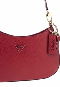 Borsa a tracolla rossa con texture, forma curva, minuteria dorata, logo triangolare e etichetta removibile. La tracolla è nera con accenti dorati.