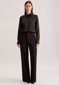Donna con lunghi capelli castani e lisci, indossa una camicia nera in satin con bottoni e pantaloni neri a vita alta e gamba larga, in piedi contro uno sfondo chiaro.