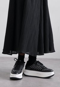 Le sneaker platform nere presentano una parte superiore in pelle liscia con inserti in occhioletto argentati, una suola bianca a contrasto e lacci neri. Una gonna nera testurizzata si adagia attorno.