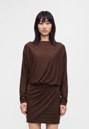 Femme aux cheveux noirs raides, portant une robe marron à manches longues avec une jupe drapée, debout devant un fond clair uni.