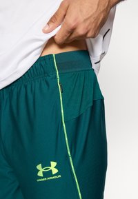 Σκούρα τιρκουάζ αθλητικά σορτς με υφή από δίχτυ, κίτρινο-πράσινες λεπτομέρειες και ελαστική μέση. Διαθέτουν μία μικρή τσέπη με φερμουάρ.