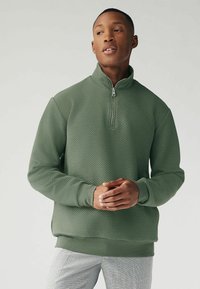 Pull-over zippé vert texturé avec un col haut, présentant un motif en losange, des poignets côtelés et des fentes latérales pour plus de liberté de mouvement.