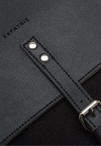 Zwarte, gestructureerde leren tas met stiksels, een metalen gesp en een logo dat op het oppervlak is geperst. Voorzien van een stevige schouderband met klinknagels.