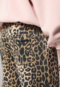 Pantalones de mezclilla con estampado de leopardo en tonos marrón y negro, que presentan un bolsillo trasero con detalle de costura y parte de un sudadera rosa arriba.