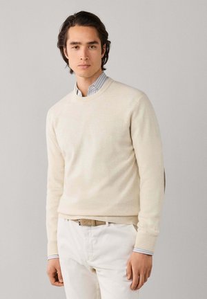 Joven con un suéter crema sobre una camisa de rayas con cuello y pantalones blancos, de pie frente a un fondo liso de color gris claro.
