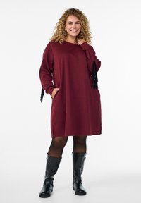 Abito felpa bordo con maniche lunghe, dettagli di frange, tasche e spacchi laterali. Abbinato a stivali neri al ginocchio, texture liscia.