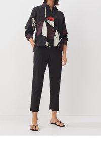 Chemise à manches courtes, boutonnée, de couleur noire avec des motifs abstraits multicolores ; associée à un pantalon noir ajusté et des tongs noires.
