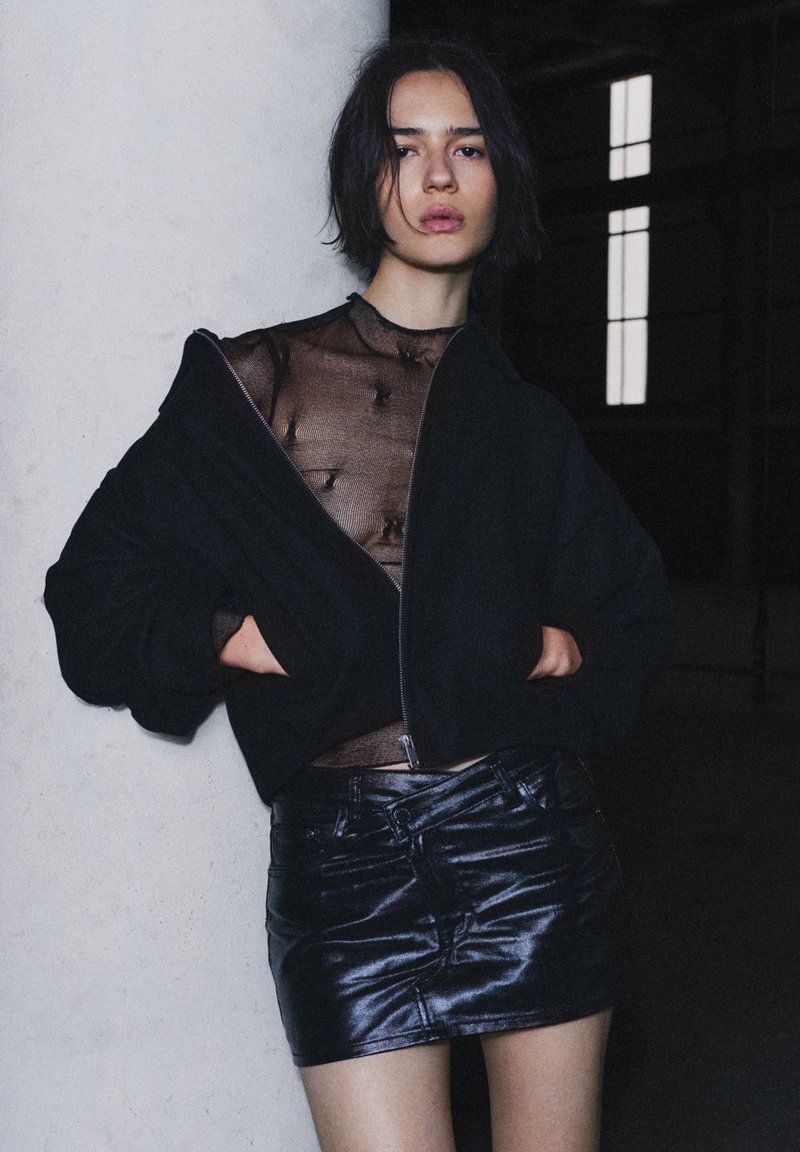 Sort bomberjakke med lynlås over en gennemsigtig top med teksturerede detaljer, kombineret med en skinnende sort læder miniskirt. Urban baggrund.