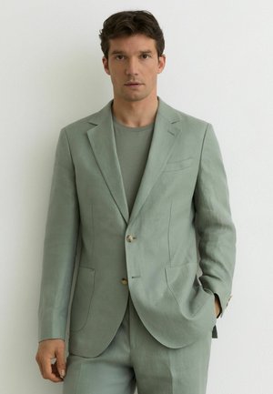 Mees, kes kannab helerohelist ülikonda ja sobivat t-särki, seisab valge seina ees, üks käsi taskus, vaadates otse ette.