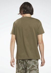 Osoba s krátkými tmavými vlasy, oblečená v olivově zeleném tričku a maskáčových kalhotách, zobrazená zezadu na jednobarevném pozadí.