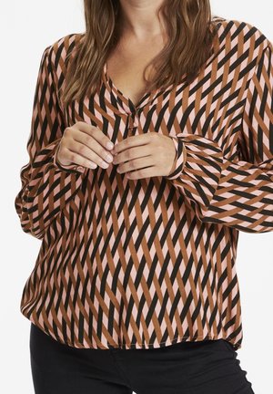 Donna che indossa una camicetta a maniche lunghe con un motivo geometrico di strisce diagonali marroni, rosa e nere, abbinata a pantaloni neri.