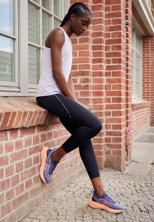 Young woman in athletic wear sitting on a brick ledge outside, wearing purple running shoes and black leggings, looking down thoughtfully.
