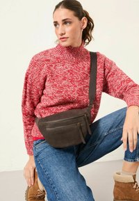 Braune Wildleder-Crossbody-Tasche mit zwei Reißverschlussfächern, getragen über einem roten Strickpullover und blauen Jeans, kombiniert mit braunen Stiefeln.