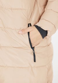 Polstret beige jakke med en lynlåslomme, elastiske håndledsmanchetter og horisontalt quiltet tekstur for isolering.