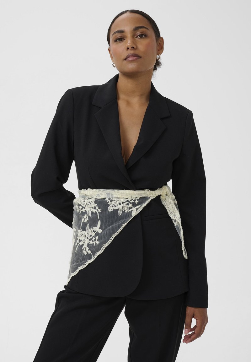 Vit bälte med spetsöverdrag och blommönster, buren över en svart skräddarsydd blazer. Slät tyg med intrikata spetsdetaljer.