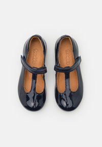 Tummansininen lakattu lasten Mary Jane -kengät, joissa on tarranauhat ja vaaleanruskeat sisäpohjat, joihin on painettu brändin nimi.