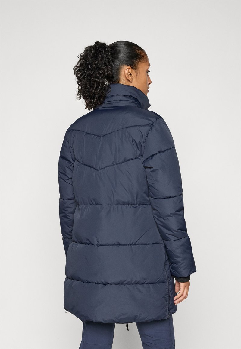 Femme avec une queue de cheval bouclée portant une veste en duvet matelassée bleu marine, de dos, sur un fond clair et uni.