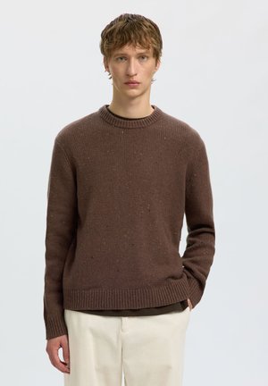 Jeune homme aux cheveux bruns courts portant un pull en maille marron et un pantalon blanc cassé, debout devant un fond clair uni.