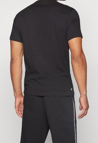 Mies, který má na sobě jednoduché černé tričko s krátkým rukávem a černé kalhoty s bílým bočním pruhem, zobrazený zezadu na jednoduchém světlém pozadí.