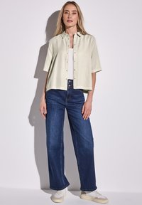Camicia verde chiaro a maniche corte con bottoni e taglio dritto, abbinata a jeans larghi blu scuro; il modello è in piedi contro uno sfondo neutro.
