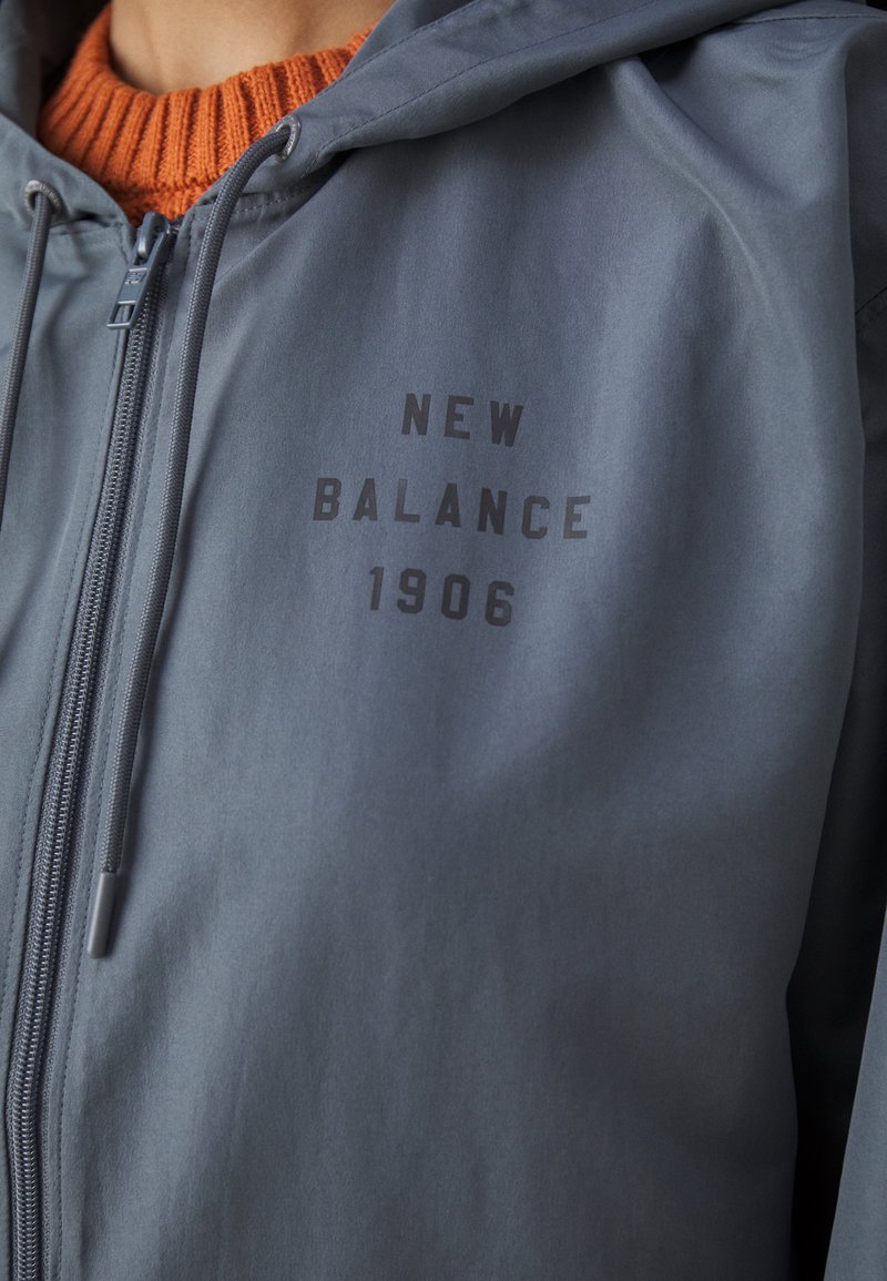 Nahaufnahme des Brustbereichs in einer blauen New Balance Jacke über einem orangefarbenen Strickpullover, der Reißverschluss ist teilweise geschlossen.