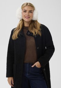 Cappotto nero con trama e due bottoni visibili, indossato sopra un maglione marrone e jeans blu; abbinato a paraorecchie bianchi e soffici.