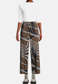 Kvinna i vida byxor med bruna, svarta och grå abstrakta virvlar, vit åtsittande topp och beige snörstövlar.