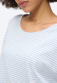 Νεαρό άτομο που φοράει μπλε και άσπρη ριγε μπλούζα με στρογγυλή λαιμόκοψη και μικρό κεντημένο λογότυπο "VD" στο στήθος.