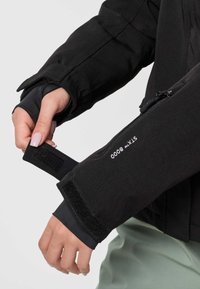 Handen die de manchetten van een zwart jasje aanpassen met een klittenbandsluiting met het label "STYX 8000,", waarbij een deel van een lichtgroene kledingstuk eronder zichtbaar is.