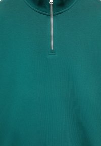 Chemise à manches longues bleu sarcelle avec une fermeture éclair argentée qui descend jusqu'à mi-hauteur à partir du col rond, montrant la texture du tissu tricoté.