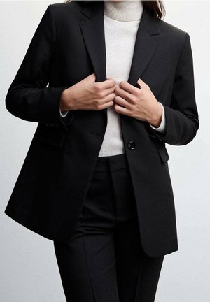 Personne ajustant un blazer noir sur un pull à col roulé blanc, portant un pantalon noir assorti, sur un fond clair uni.