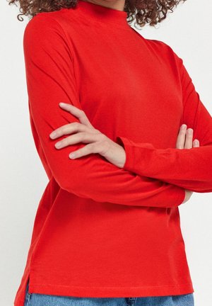 Osoba o kręconych włosach, ubrana w czerwoną bluzkę z długim rękawem i niebieskie dżinsy, stoi z założonymi ramionami na tle jednolitego tła.