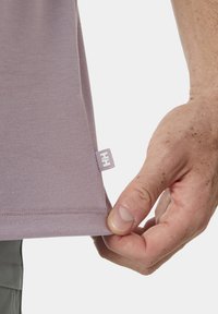 Camiseta de color púrpura claro hecha de una tela suave. Presenta una etiqueta lateral con el logotipo, costuras cuidadas y un ajuste relajado en el dobladillo. Mano sujetando el dobladillo.