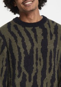 Pull en maille surdimensionné de couleur vert olive avec des motifs abstraits bleu marine foncé, col rond et tissu texturé. Design décontracté adapté pour le superposition.