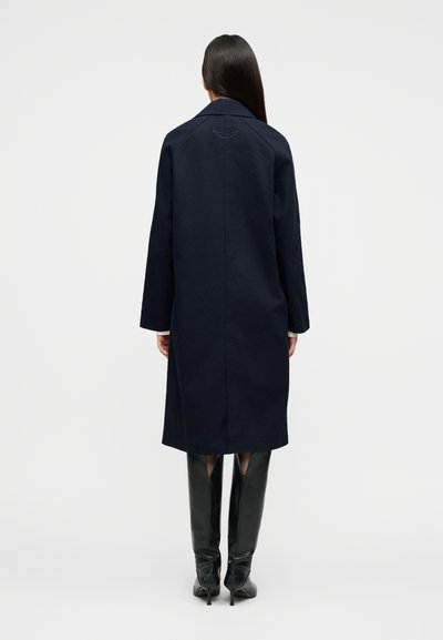 Γυναίκα με μακριά σκούρα μαλλιά που φορά παλτό μήκους μέχρι το γόνατο σε ναυτικό μπλε χρώμα και μαύρες μπότες με ψηλό τακούνι, γυρισμένη με την πλάτη σε ένα απλό φόντο.