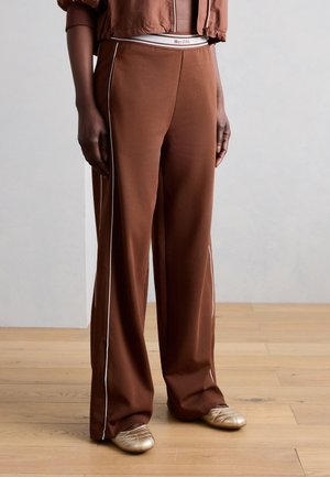 Άτομο που φοράει καφέ παντελόνια με φαρδιά λάστιχα και λευκές πλαϊνές ρίγες, χρυσά καπιτονέ παπούτσια slip-on και cropped καφέ μπουφάν σε ξύλινο πάτωμα.