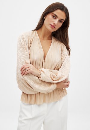 Blouse plissée beige pâle avec un décolleté en V profond, des manches ballon longues et des poignets froncés, terminée par un ourlet volanté.