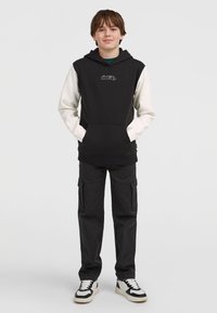 Čierno-biela mikina s predným vreckom, paired with �čierne cargo nohavice. Mikina má vyšitú nášivku a nohavice majú zipsové detaily.