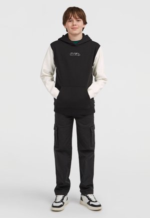 Čierno-biela mikina s predným vreckom, paired with čierne cargo nohavice. Mikina má vyšitú nášivku a nohavice majú zipsové detaily.