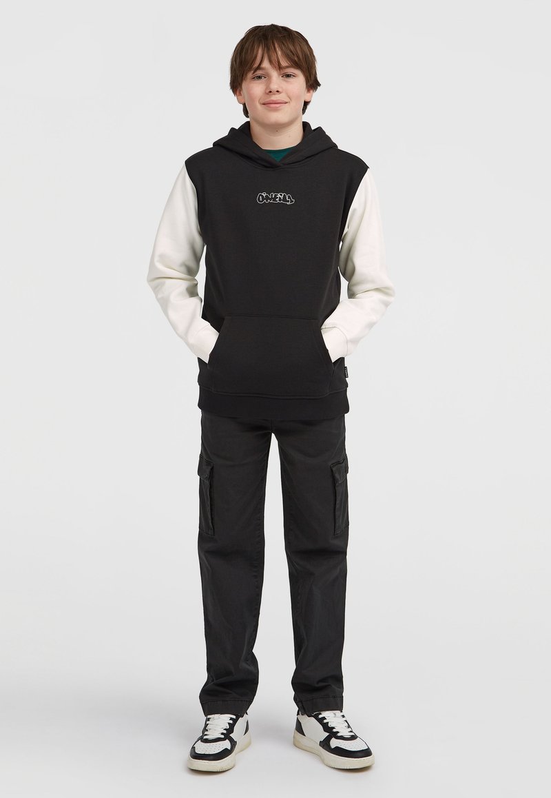 Čierno-biela mikina s predným vreckom, paired with čierne cargo nohavice. Mikina má vyšitú nášivku a nohavice majú zipsové detaily.