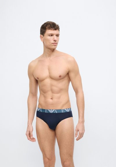 Άνδρας χωρίς μπλούζα με κοντά σγουρά μαλλιά, φορώντας μπλε σκούρο εσώρουχο με μπλε λογότυπο στη μέση, στέκεται μπροστά σε απλό λευκό φόντο.