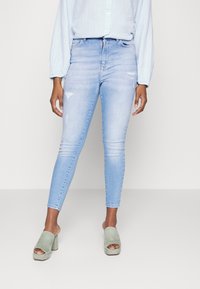 Jeans skinny azul claro com detalhes desgastados, cintura alta e fecho de zíper, combinados com sandálias de bloco de camurça verde de abertura na ponta.