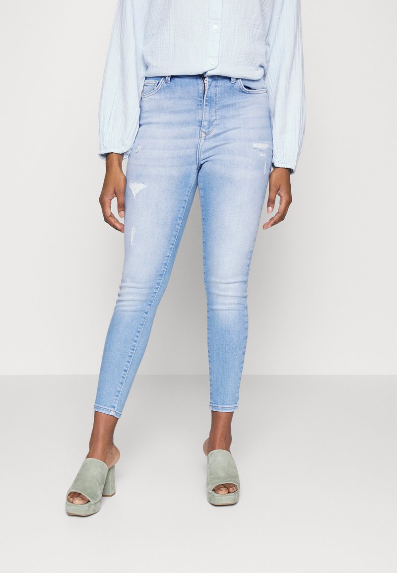 Jeans skinny azul claro com detalhes desgastados, cintura alta e fecho de zíper, combinados com sandálias de bloco de camurça verde de abertura na ponta.