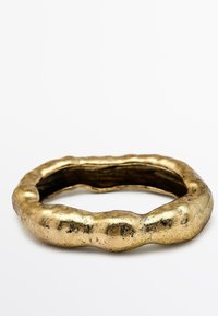 Goudkleurige metalen armband met een gladde, onregelmatige vorm, voorzien van een gestructureerd, bobbelig oppervlak en een holle binnenkant.