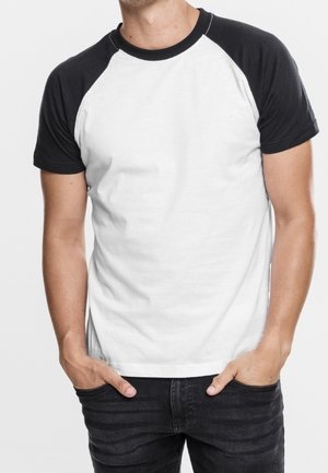 Mand iført en hvid t-shirt med sorte raglanærmer og sorte jeans, hænderne i lommerne, stående mod en ensfarvet baggrund.