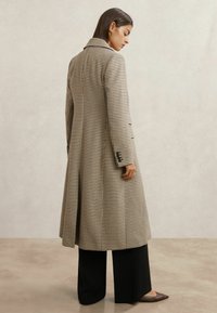 Kvinde iført en lang frakke med hundetandsmønster, sorte bukser med brede ben og brune spidse sko, stående og kiggende nedad til siden.