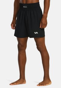 Man in zwarte RVCA-shorts die blootsvoets tegen een effen achtergrond staat met ontspannen handen langs de zij.