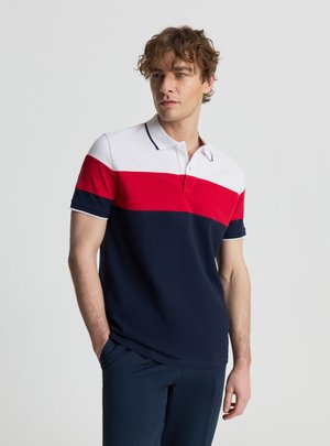 Uomo che indossa una polo a maniche corte con blocchi di colore orizzontali bianco, rosso e blu navy, in piedi con le mani nelle tasche, che guarda di lato.