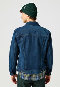 Chaqueta de mezclilla azul oscura con un corte clásico, puños abotonados y herrajes de cobre. Lleva puesta una camisa de cuadros en verde y azul, mostrando un estilo en capas.
