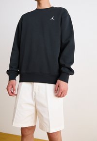 Sudadera negra de manga larga, con puños y cuello de canalé. Presenta un pequeño logo blanco de Jordan en el pecho, combinado con pantalones cortos blancos.